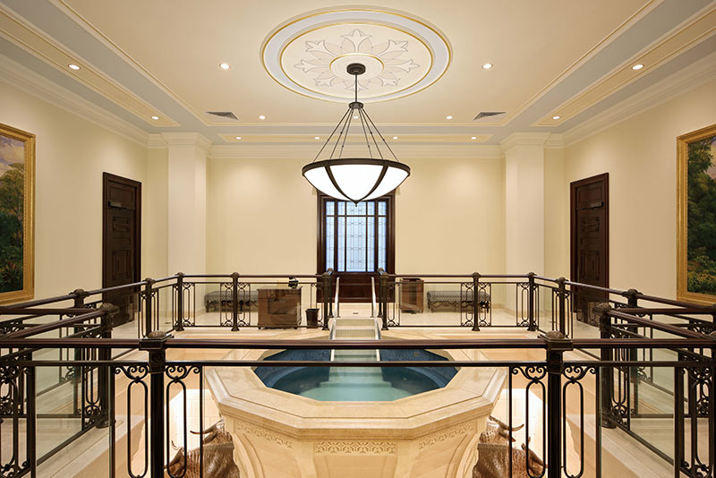 Interior photo of the Baptistry at the Latter-Day Saints temple. A large limestone pool centers the room with bronze railing surrounding.