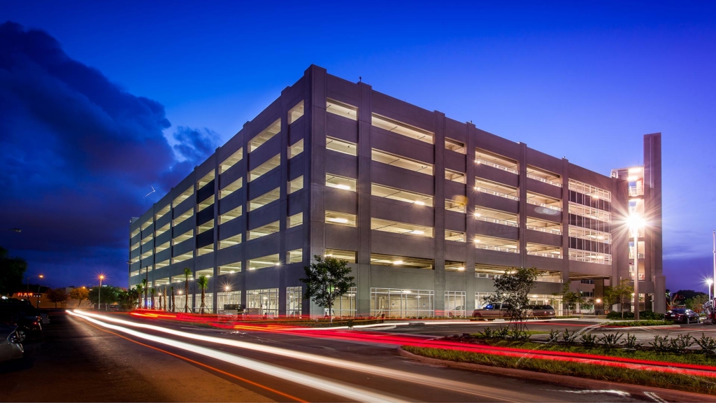 Exterior photo of the Miami Dade College Hialeah Campus parking garage. Evening photo with bright blue skies. Garage lights on. Vehicle lights create streaks on the adjacent street.