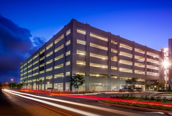 Exterior photo of the Miami Dade College Hialeah Campus parking garage. Evening photo with bright blue skies. Garage lights on. Vehicle lights create streaks on the adjacent street.