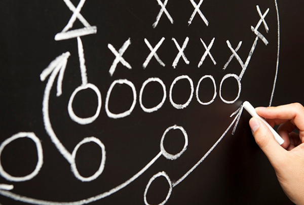 Hand diagramming football play in chalk on chalkboard.