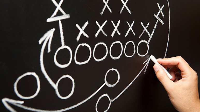 Hand diagramming football play in chalk on chalkboard.