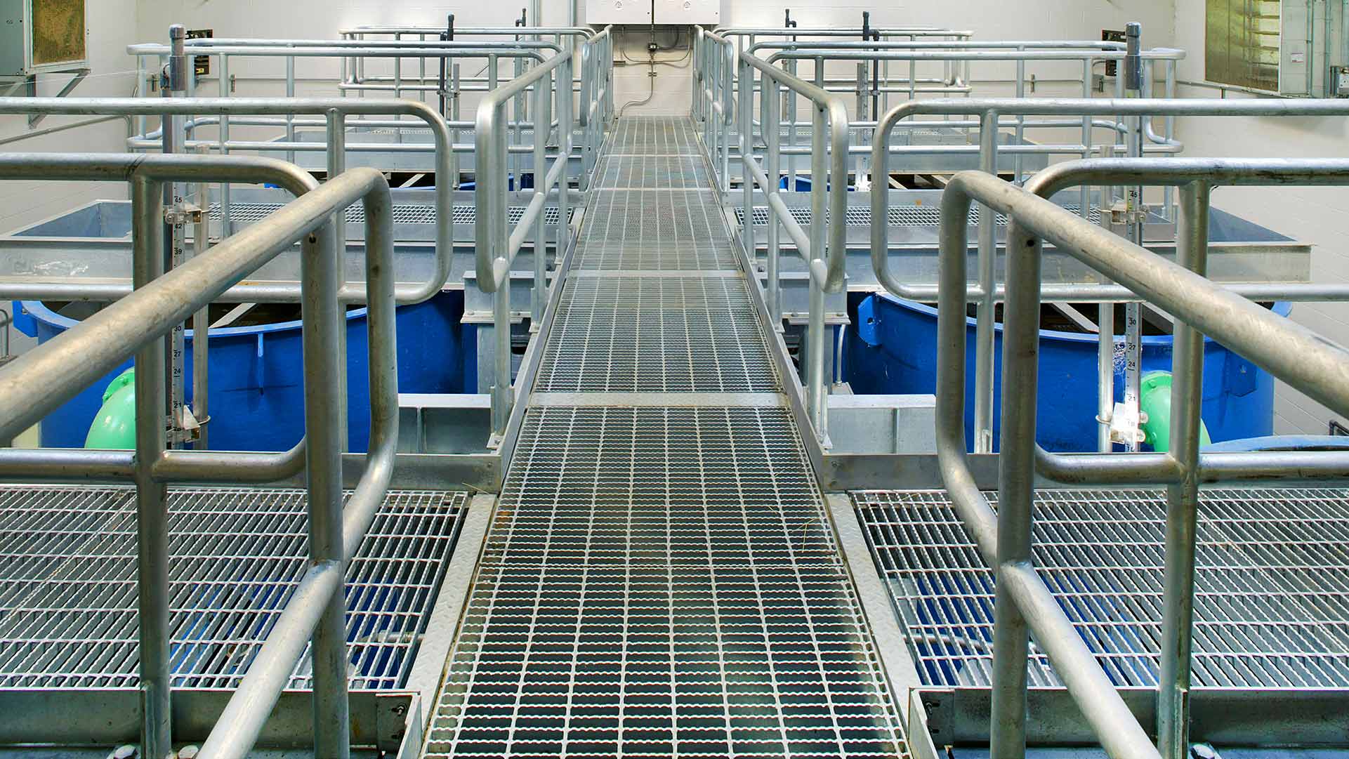 Aluminum grating walkways above six large blue tanks at the Indian Head Wastewater Treatment Plant.|Aluminum grating walkways above six large blue tanks with green piping at the Indian Head Wastewater Treatment Plant.|Exterior photo looking up at 2 story red brick facility. Exterior stairs let up to the second floor.|Exterior photo of the Indian Head Wastewater Treatment Plant|Large green and grey pipes with electrical and mechanical valves rest on concrete blocks inside the facility.|||||