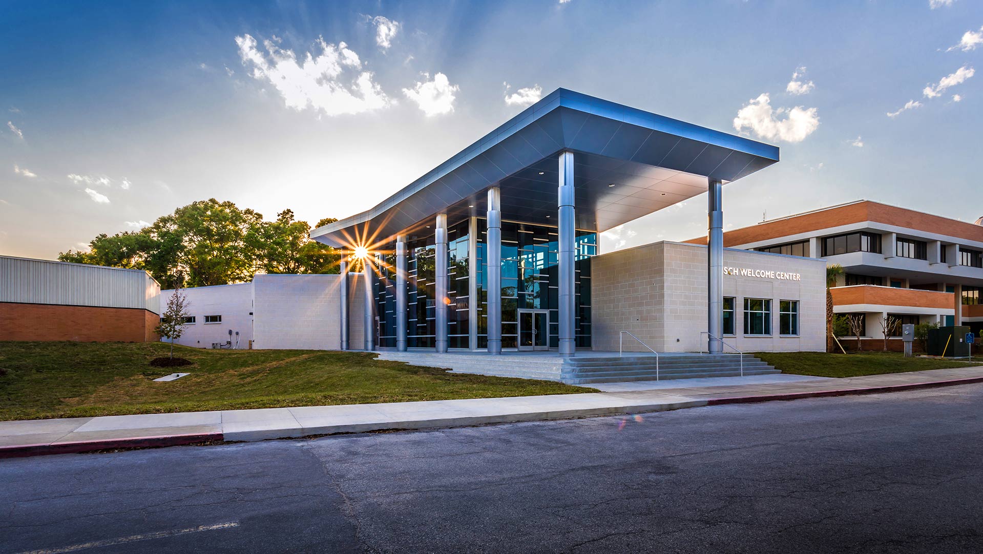 Sun setting behind the Frisch Welcome Center|Frisch Welcome Center exterior|Frisch Welcome Center doorway with view of grounds|Frisch Welcome Center offices|Frisch Welcome Center exterior|Frisch Welcome Center entry area with logo inset in terrazzo floor|Frisch Welcome Center exterior at dusk|Frisch Welcome Center group presentation space|Frisch Welcome Center lobby featuring Jacksonville University logo inset in terrazzo floor|Education Blue Background||||||||||