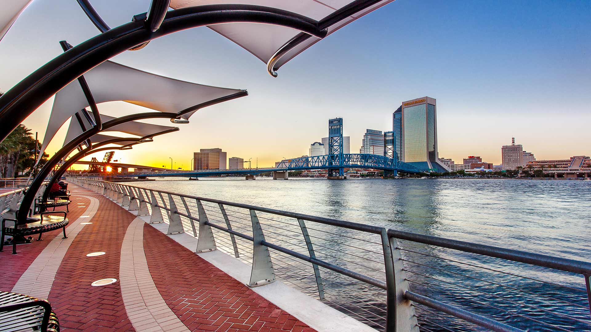 Photo of Jacksonville's Southbank Riverwalk at sunset. Red brick sidewalk alongside the river with steel guardrails. Canopies overhang the sidewalks. Jacksonville skyline in background.