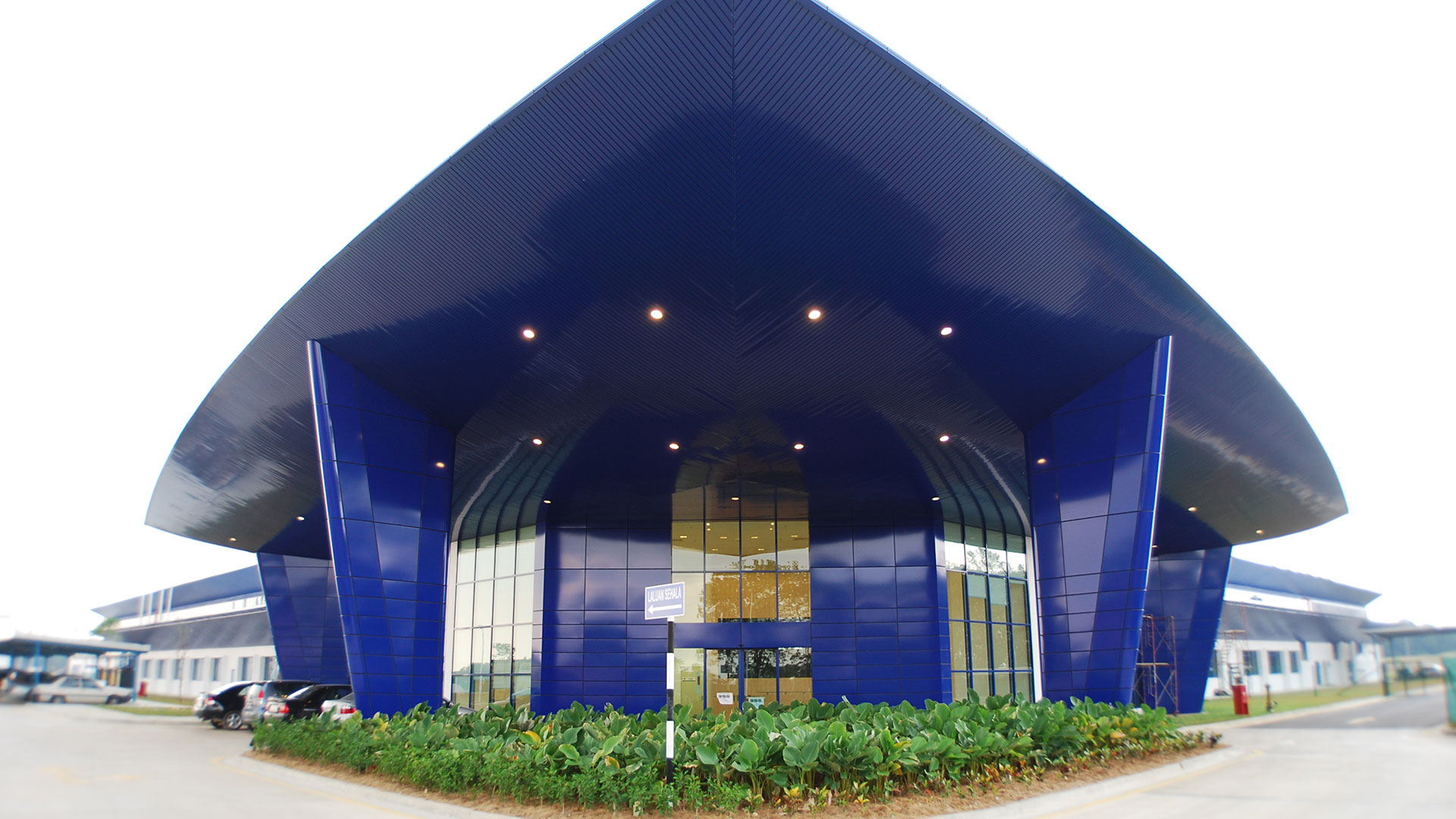 Exterior entrance of Spirit AeroSystems SE Asia. Bright blue airfoil inspired triangular building.|Aviation Blue Background|Exterior entrance of Spirit AeroSystems SE Asia. Bright blue triangular building.|Reception desk at Spirit AeroSystems SE Asia.|Interior of Spirit AeroSystems SE Asia. Large industrial open room with yellow railing and florescent lighting.|Advanced Manufacturing Blue Background Rolls Royce|||||