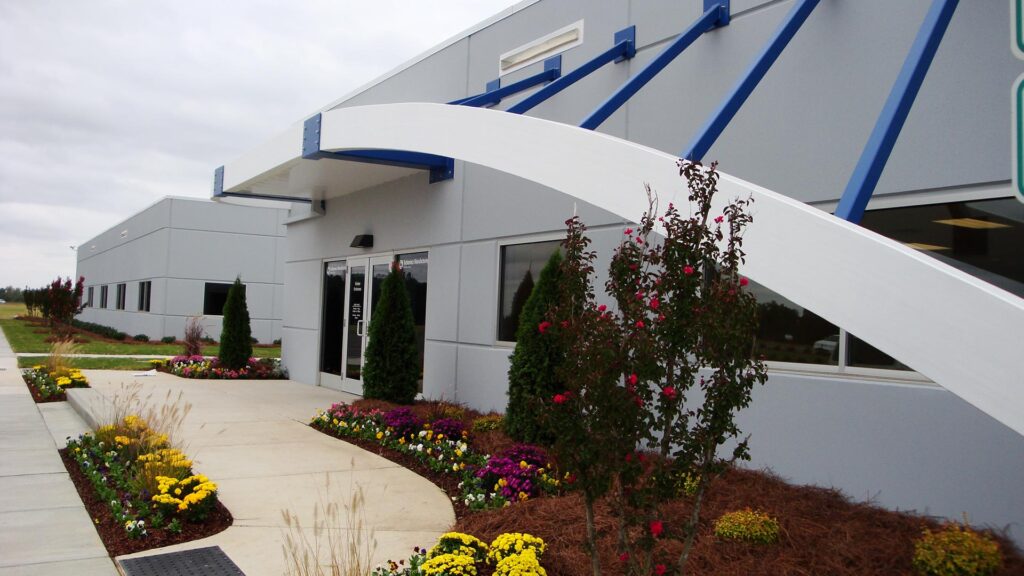 Front entrance of Turbomeca TMM Manufacturing Facility. Awning supported by blue structures. Floral landscaping.|