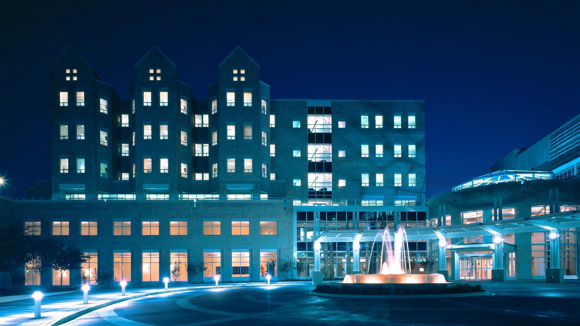 Exterior photo of Wolfson Children's Hospital at night. Blue lights and fountain surrounded by brick circle drive.|Interior photo of main entry. Glass dome and full height windows on the walls surround main doors.|Interior photo of children's play area. Glass windows and door separate room from hallway|Exterior photo of Wolfson Children's Hospital during the day. Brick drive leads up to six-story tan bricked building.|Interior photo of hospital room. Crib bed and chair sit in the middle of the room.|||||