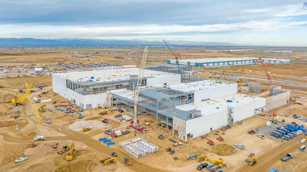Aerial view of a large manufacturing facility under construction with cranes, structural steel, and heavy equipment.