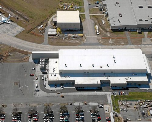 Aerial view of Gulfstream Paint Hangar. White building surrounded by vehicle parking lots and aircraft parked on apron.