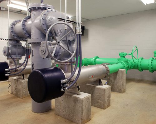 Large green and grey pipes with electrical and mechanical valves rest on concrete blocks inside the facility.