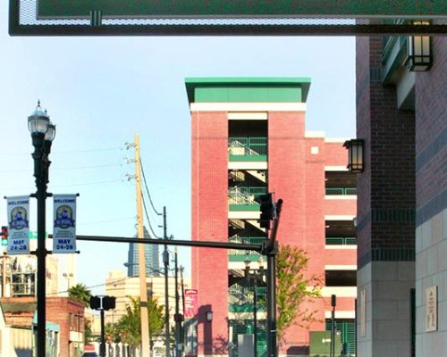 Exterior photo of Jacksonville Municipal Garages. Photo is through a doorway outside the arena. Trees line the street. Red brick with green trim accent the garage. Sign above doorway reads 'Exit Home Plate Gate'.