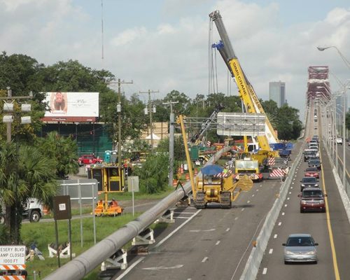 Very long pipe on stands along a highway full of cars. Large tractors, a crane, and other equipment there for the installation.