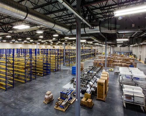 Interior of large warehouse. Empty shelving racks occupy half the room. Boxes of supplies are on pallets on the floor occupy the other half of the room.