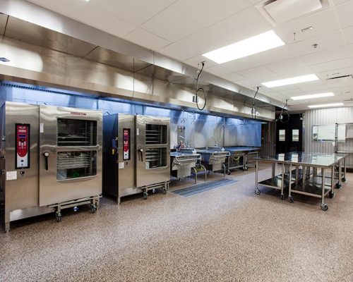 Interior photo of large room with stainless steel equipment: tables, sinks, shelving, ovens.