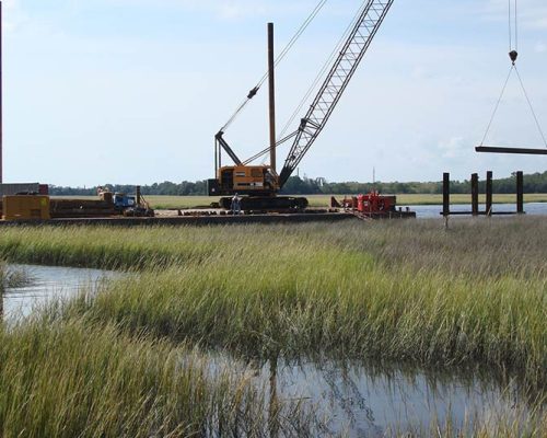 A crane lifts a beam across a swampy grassy marsh.