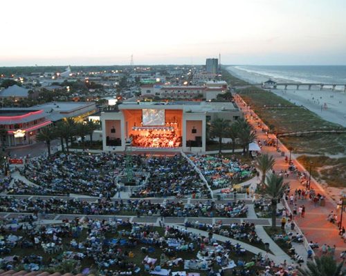 Birds-eye view of large crowd gathered around the Sea Walk Pavilion listening to a concert at dusk.