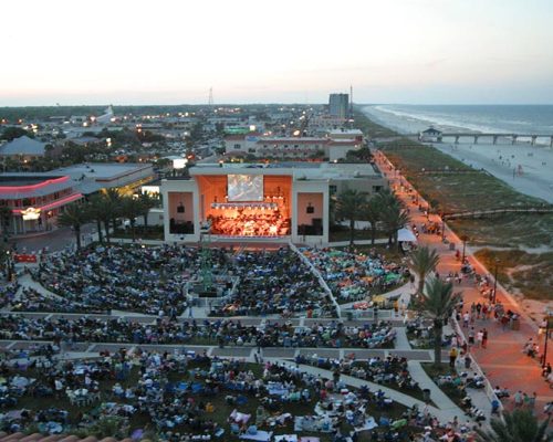 Birds-eye view of large crowd gathered around the Sea Walk Pavilion listening to a concert at dusk.