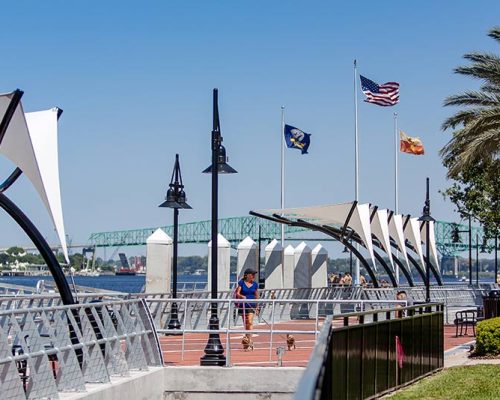 Photo of Jacksonville's Southbank Riverwalk during a sunny day. Brick sidewalk alongside the river with steel guardrails. Canopies overhang the sidewalk providing shade.