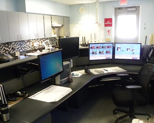 Interior photo of a workstation with 3 monitors. Upper cabinets and exit door are in the background.