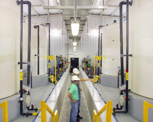 Interior photo of the facility with large white tanks. Men in hardhats checking their status.